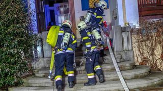 Drei Feuerwehrleute, die Helme tragen und Ausrüstung mit sich führen, erklimmen die Treppe, um ein Gebäude zu betreten. Sie haben Sauerstoffflaschen und einen Feuerlöscher bei sich.