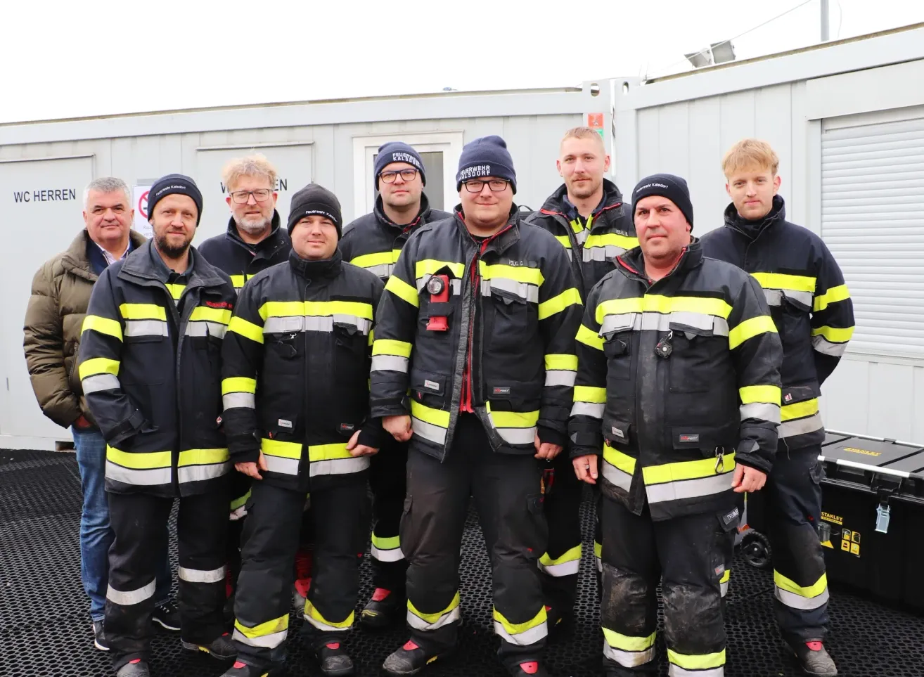 Eine Gruppe von Feuerwehrleuten in schwarzen und gelben Uniformen steht vor einem Gebäude. Sie tragen alle Mützen und Brillen. Das Gebäude hat eine weiße Wand und eine Tür.
