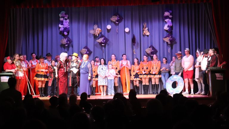 Eine Gruppe von Menschen steht auf der Bühne, einige in Kostümen, möglicherweise bei einem Schulstück. Die Bühne hat einen blauen Vorhang und dekorative Aufhängungen.