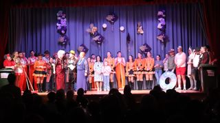 Eine Gruppe von Menschen steht auf der Bühne, einige in Kostümen, möglicherweise bei einem Schulstück. Die Bühne hat einen blauen Vorhang und dekorative Aufhängungen.