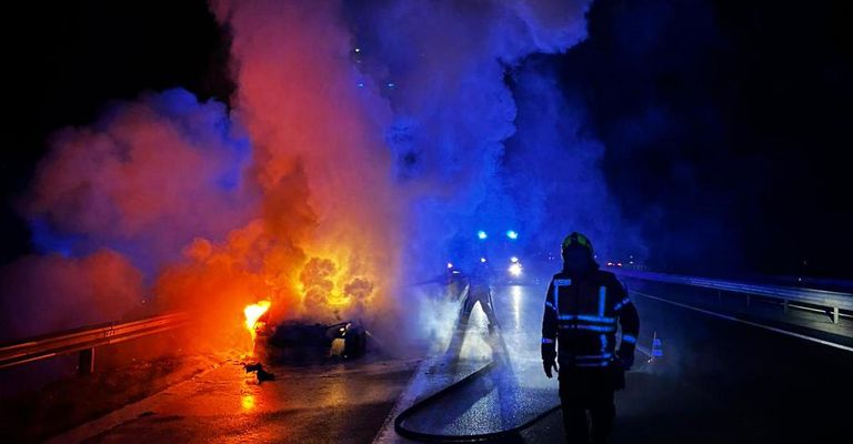 Ein Auto brennt nachts am Straßenrand, Feuerwehrleute und Polizei sind vor Ort.
