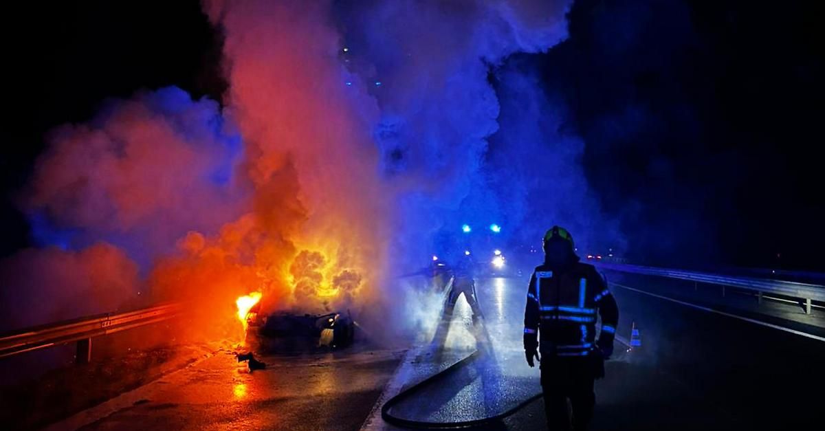 Ein Auto brennt nachts am Straßenrand, Feuerwehrleute und Polizei sind vor Ort.
