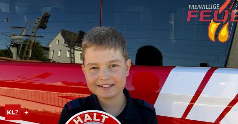 Ein junger Junge steht vor einem roten Feuerwehrauto, lächelt und hält ein kreisförmiges Schild mit der Aufschrift 'HALT'. Hinter ihm ist ein Fenster mit der Reflexion eines Hauses und eines Baumes.