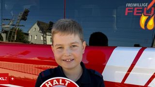 Ein junger Junge steht vor einem roten Feuerwehrauto, lächelt und hält ein kreisförmiges Schild mit der Aufschrift 'HALT'. Hinter ihm ist ein Fenster mit der Reflexion eines Hauses und eines Baumes.