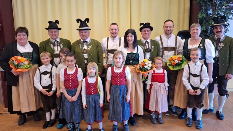 Eine Gruppe von Menschen, einschließlich Erwachsener und Kindern, posiert für ein Foto. Sie sind in traditioneller Kleidung gekleidet, wobei die Frauen Blumenkränze tragen und die Kinder Blumen halten. Der Hintergrund scheint drinnen zu sein, mit einem gelben Vorhang.