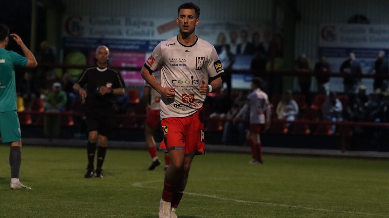 Ein Fußballer läuft mit erhobenem Daumen auf dem Spielfeld. Im Hintergrund befinden sich mehrere andere Spieler und Zuschauer.