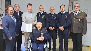 Ein älterer Mann im Rollstuhl erhält eine Auszeichnung, während ihn uniformierte Offiziere umgeben. Sie stehen in einem Raum mit einer weißen Wand.