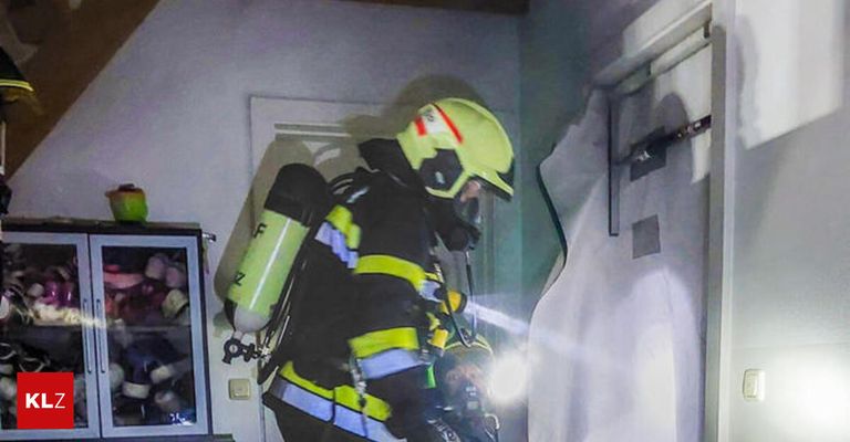 Ein Feuerwehrmann mit gelbem Helm und Sauerstofftank steht in einem Raum mit beschädigter Wand. Ein weiterer Feuerwehrmann ist hinter ihm.