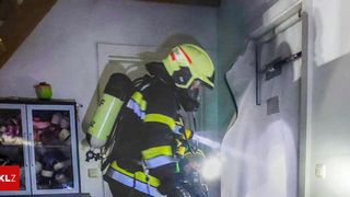 Ein Feuerwehrmann mit gelbem Helm und Sauerstofftank steht in einem Raum mit beschädigter Wand. Ein weiterer Feuerwehrmann ist hinter ihm.