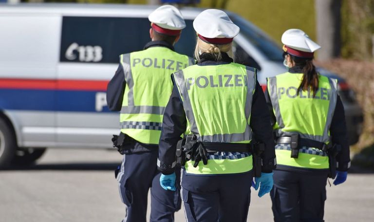 Drei Polizeibeamte in reflektierenden Westen und Handschuhen gehen vor einem Polizeiwagen.