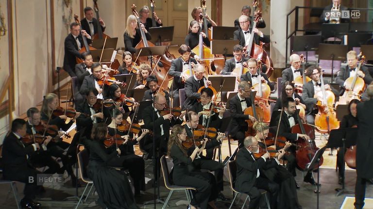Ein Symphonieorchester in formeller Kleidung spielt auf der Bühne, mit Musikern, die Violinen, Celli und andere Instrumente spielen, mit Notenständern vor ihnen.