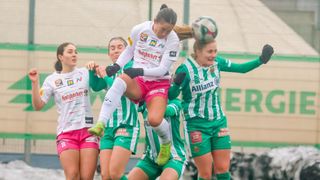 Eine Fußballspielerin in einem weißen Trikot mit pinken Shorts und grünen Schuhen springt, um den Ball zu köpfen. Sie wird von Teamkolleginnen in grünen Trikots umgeben.