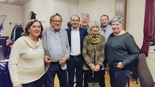 Sechs Menschen lächeln und posieren für ein Foto, mit zwei Frauen links und vier Männern in Anzügen rechts.