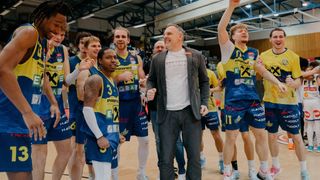 Eine Gruppe von Basketballspielern feiert auf dem Spielfeld mit einem Trainer in einer grauen Jacke.