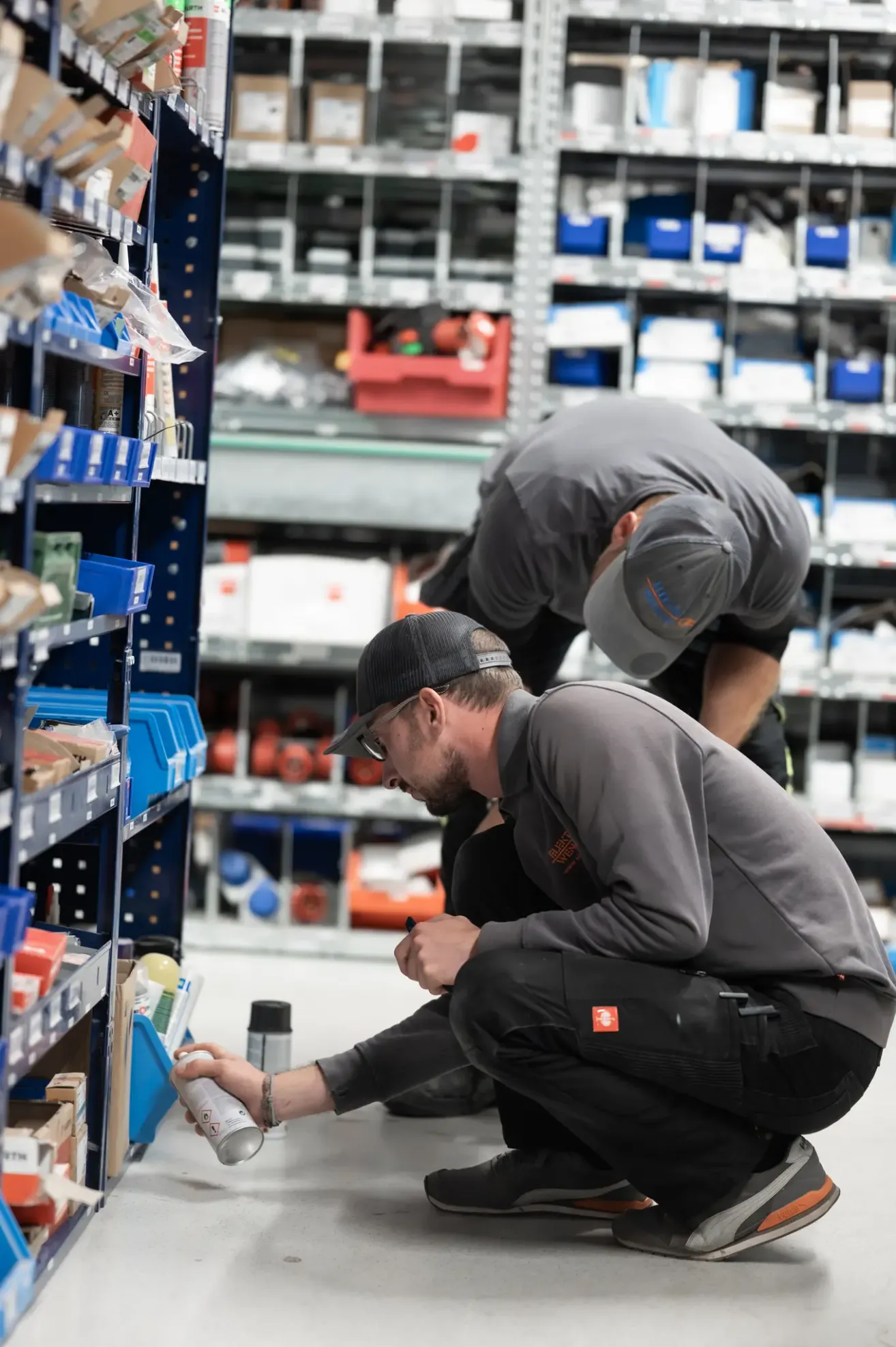Zwei Männer in grauen Uniformen befinden sich in einem Lager, einer schreibt auf ein Papier und der andere schaut sich Artikel in einem Regal an.