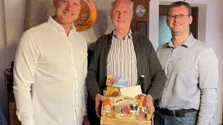 Drei Männer stehen in einem Raum, lächeln und posieren für ein Foto. Der Mann in der Mitte hält eine Holzkiste mit Essen und einer Flasche.