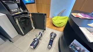 Ein beschädigter Computerbildschirm liegt offen mit freiliegenden Komponenten. In der Nähe liegen zwei schwarze und silberne Werkzeuge, beschriftet mit 'ZURDOS', auf dem Boden. Ein gelber Beanbag-Stuhl und ein blauer Stuhl sind im Hintergrund.