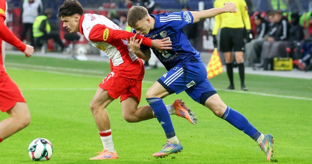 Zwei Fußballspieler in roten und blauen Trikots sind auf einem Feld in einen körperlichen Wettkampf verwickelt.