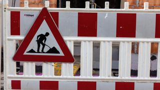 Ein rotes und weißes Dreiecksschild mit einer Silhouette einer Person beim Graben ist an einer weiß-rot gestreiften Barriere befestigt. Das Schild warnt vor bevorstehenden Straßenarbeiten.
