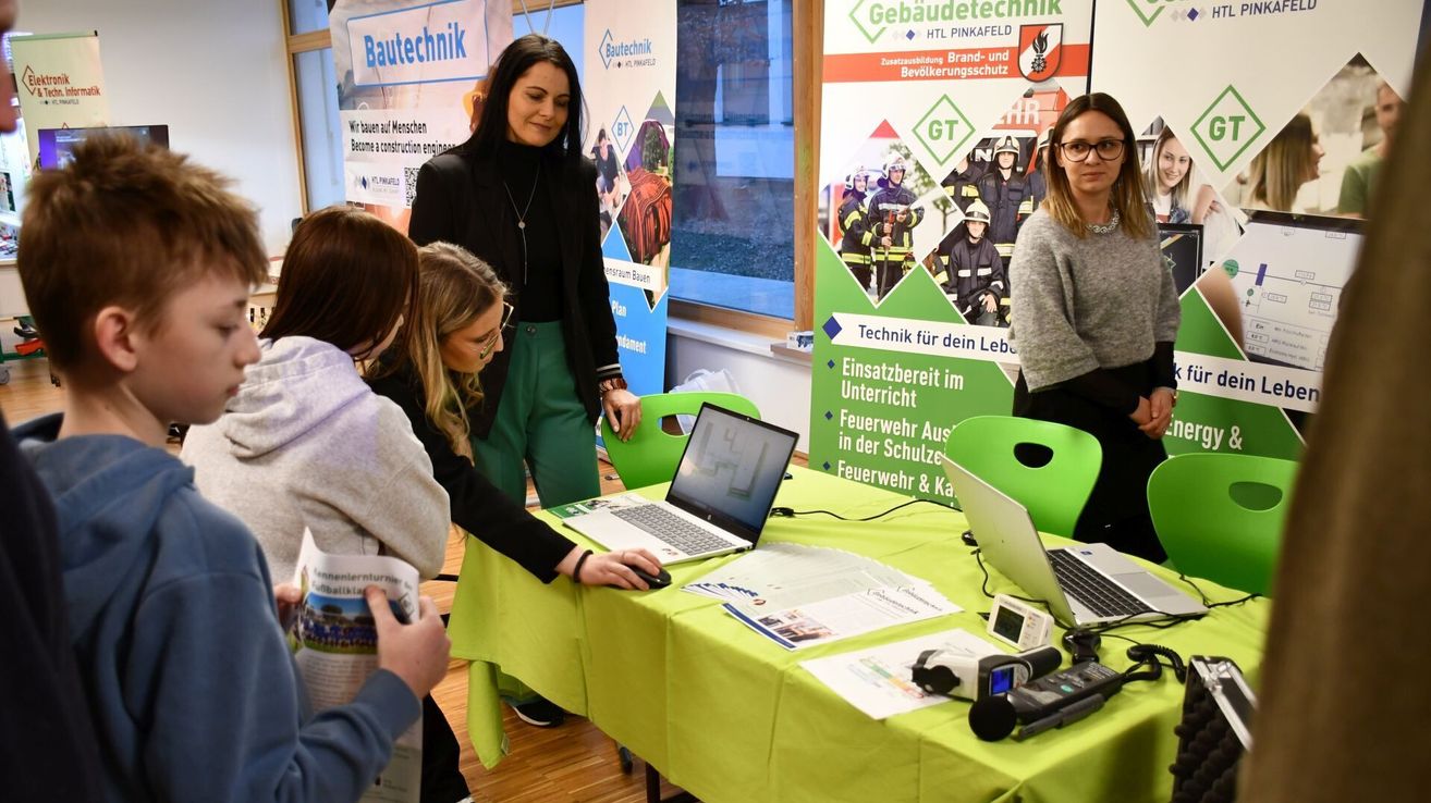 Menschen sind bei einer Veranstaltung mit einem Tisch voller Laptops, Papiere und Mikrofone. Eine Frau steht hinter ihnen, lächelt, in der Nähe eines Plakats über Gebäudetechnik.