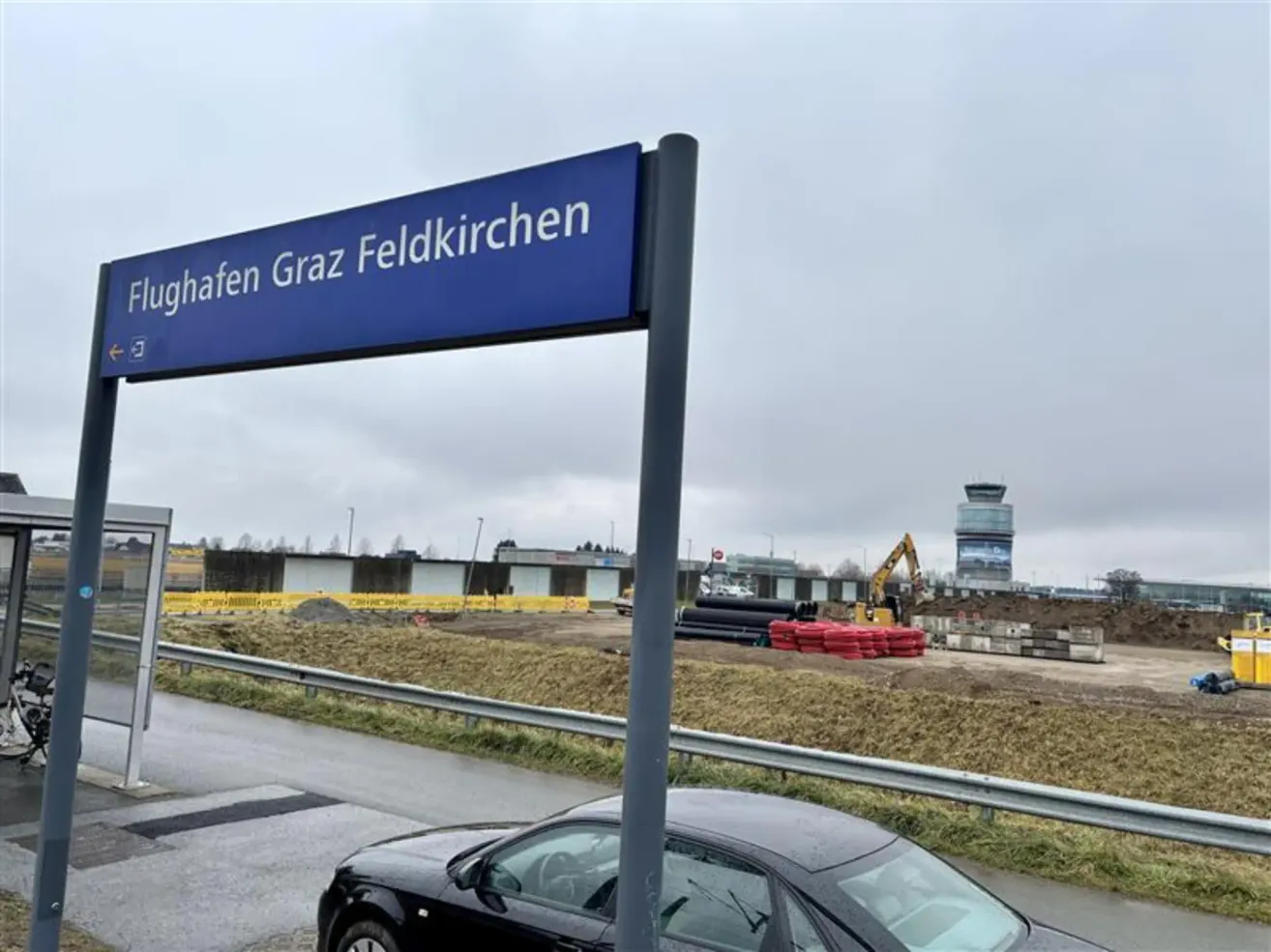 Ein blaues Straßenschild mit dem Text 'Flughafen Graz Feldkirchen' ist an einem Pfosten montiert. Ein Auto ist auf der Straße geparkt. Im Hintergrund sind Bauarbeiten im Gange.