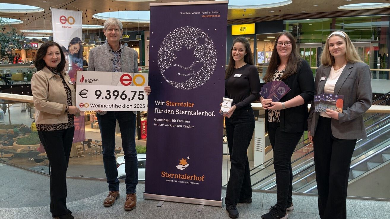Drei Personen stehen in einem Einkaufszentrum, eine hält einen großen Scheck. Hinter ihnen befindet sich ein lilafarbener Banner mit 'Sterntaler' in weiß. Zwei Frauen halten Gegenstände und eine lächelt. Im Hintergrund sind Geschäfte und Rolltreppen zu sehen.