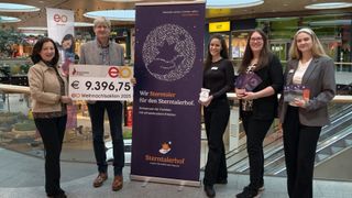Drei Personen stehen in einem Einkaufszentrum, eine hält einen großen Scheck. Hinter ihnen befindet sich ein lilafarbener Banner mit 'Sterntaler' in weiß. Zwei Frauen halten Gegenstände und eine lächelt. Im Hintergrund sind Geschäfte und Rolltreppen zu sehen.