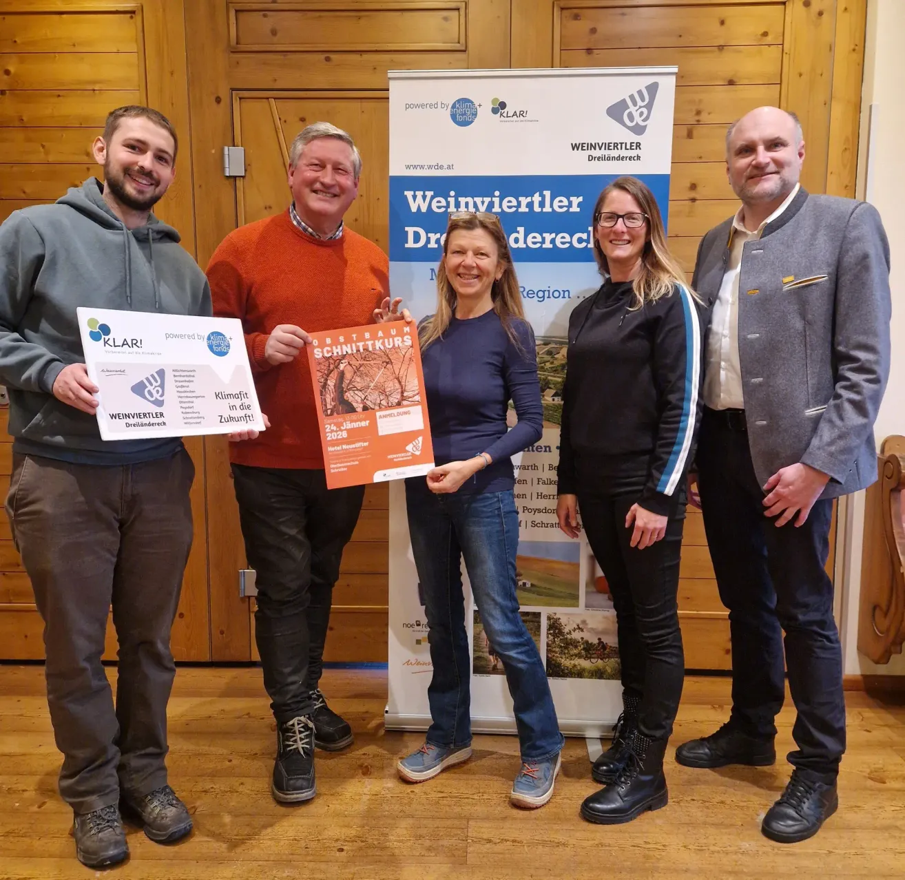 Vier Menschen stehen vor einer Holzwand und lächeln für ein Foto. Zwei Männer halten Urkunden, eine Frau ein Buch und eine andere lächelt. Hinter ihnen steht ein Banner mit der Aufschrift 'Weinviertler Dreiländereck'.