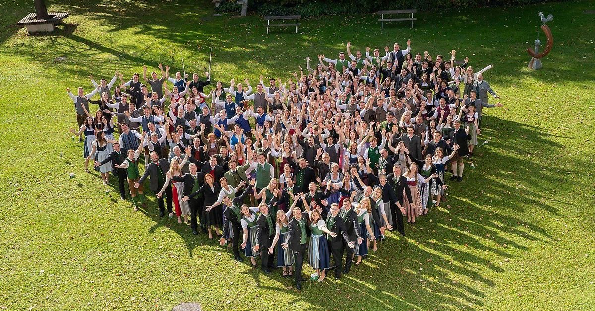 Eine große Gruppe von Menschen in traditioneller deutscher Kleidung, die auf einem Rasenfeld ein Herz bilden, wobei einige Personen die Hände heben und andere Gegenstände halten.