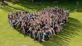 Eine große Gruppe von Menschen in traditioneller deutscher Kleidung, die auf einem Rasenfeld ein Herz bilden, wobei einige Personen die Hände heben und andere Gegenstände halten.