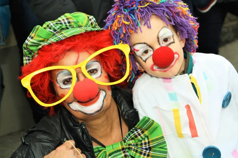 Zwei Personen, die als Clowns gekleidet sind, eine trägt gelbe Brille und einen karierten Hut, die andere hat eine bunte Perücke und eine rote Nase. Sie tragen beide weißes Gesichtsmake-up und scheinen für ein Foto zu posieren.