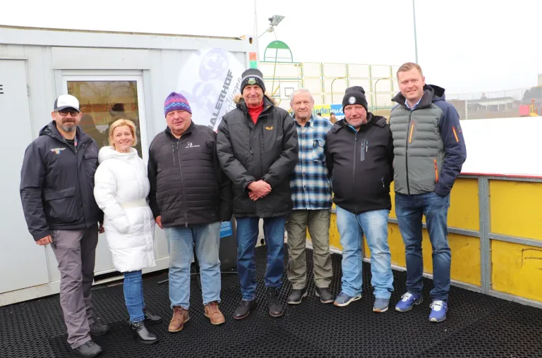 Eine Gruppe von fünf Personen steht draußen in einer kalten Umgebung, alle warm gekleidet. Zwei Männer tragen Mützen, und zwei andere tragen Mützen. Sie scheinen für ein Foto zu posieren. Hinter ihnen steht ein weißes Gebäude mit einer Glastür.