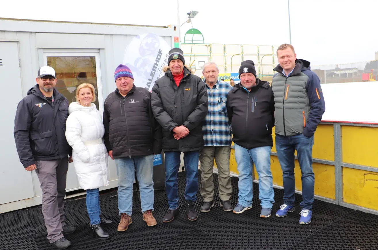 Eine Gruppe von fünf Personen steht draußen in einer kalten Umgebung, alle warm gekleidet. Zwei Männer tragen Mützen, und zwei andere tragen Mützen. Sie scheinen für ein Foto zu posieren. Hinter ihnen steht ein weißes Gebäude mit einer Glastür.