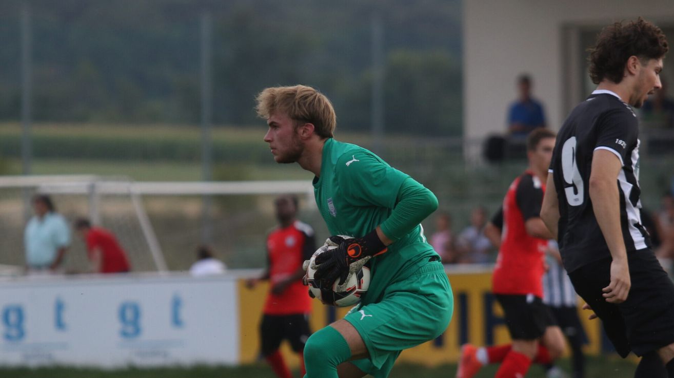 Ein Fußballspieler in grüner Uniform, mit einem Fußball in der Hand, steht auf einem Feld mit verschwommenen Spielern und Zuschauern im Hintergrund.