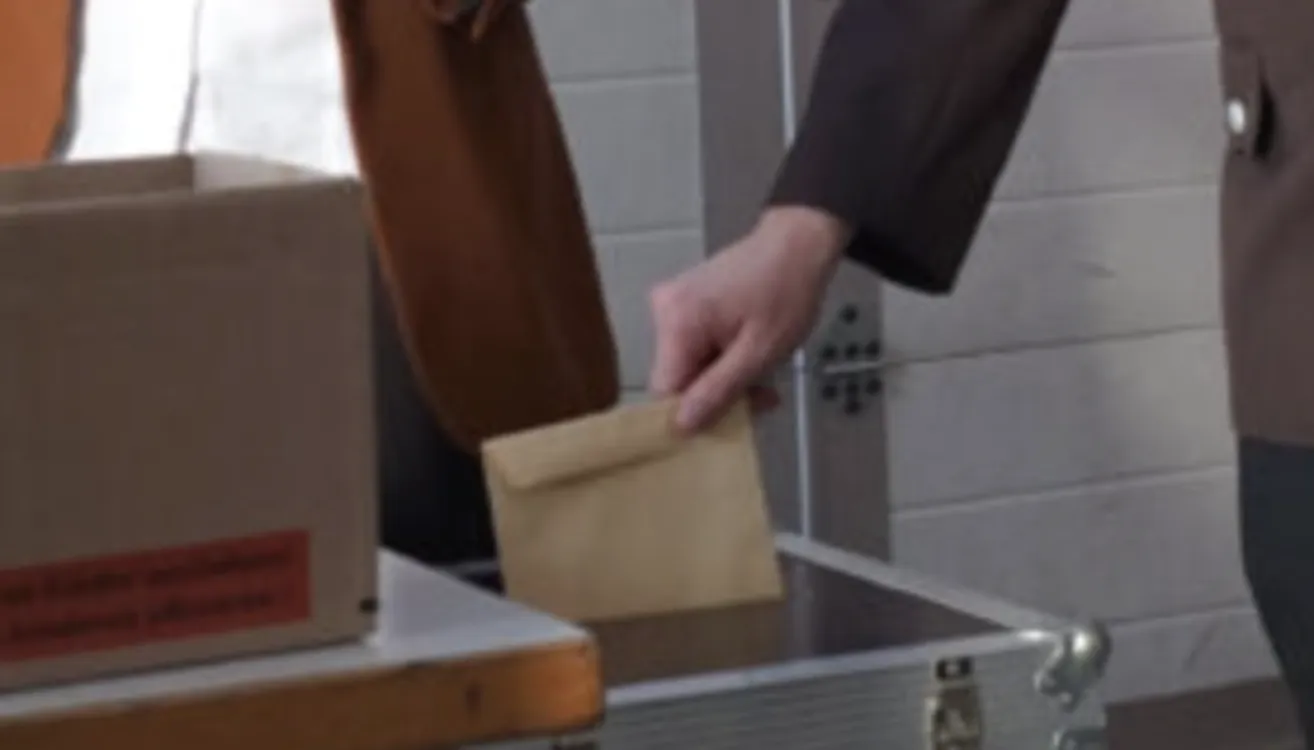 Eine Person legt einen braunen Umschlag auf eine Metalloberfläche, mit einem Kasten links und einer weißen Wand dahinter.