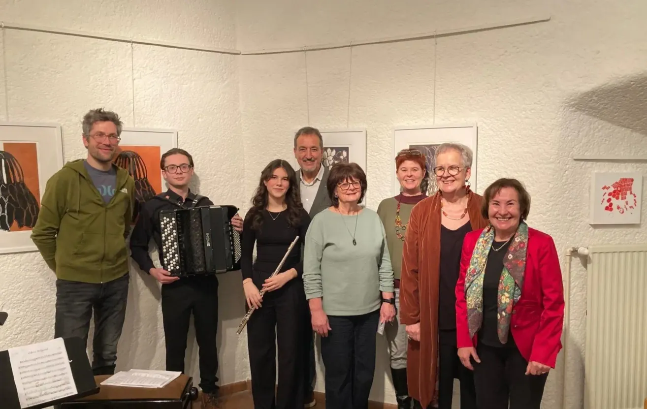 Eine Gruppe von Menschen in einem Raum mit Musikinstrumenten, lächelnd und posierend für ein Foto. Der Raum ist weiß mit gerahmten Bildern an der Wand.