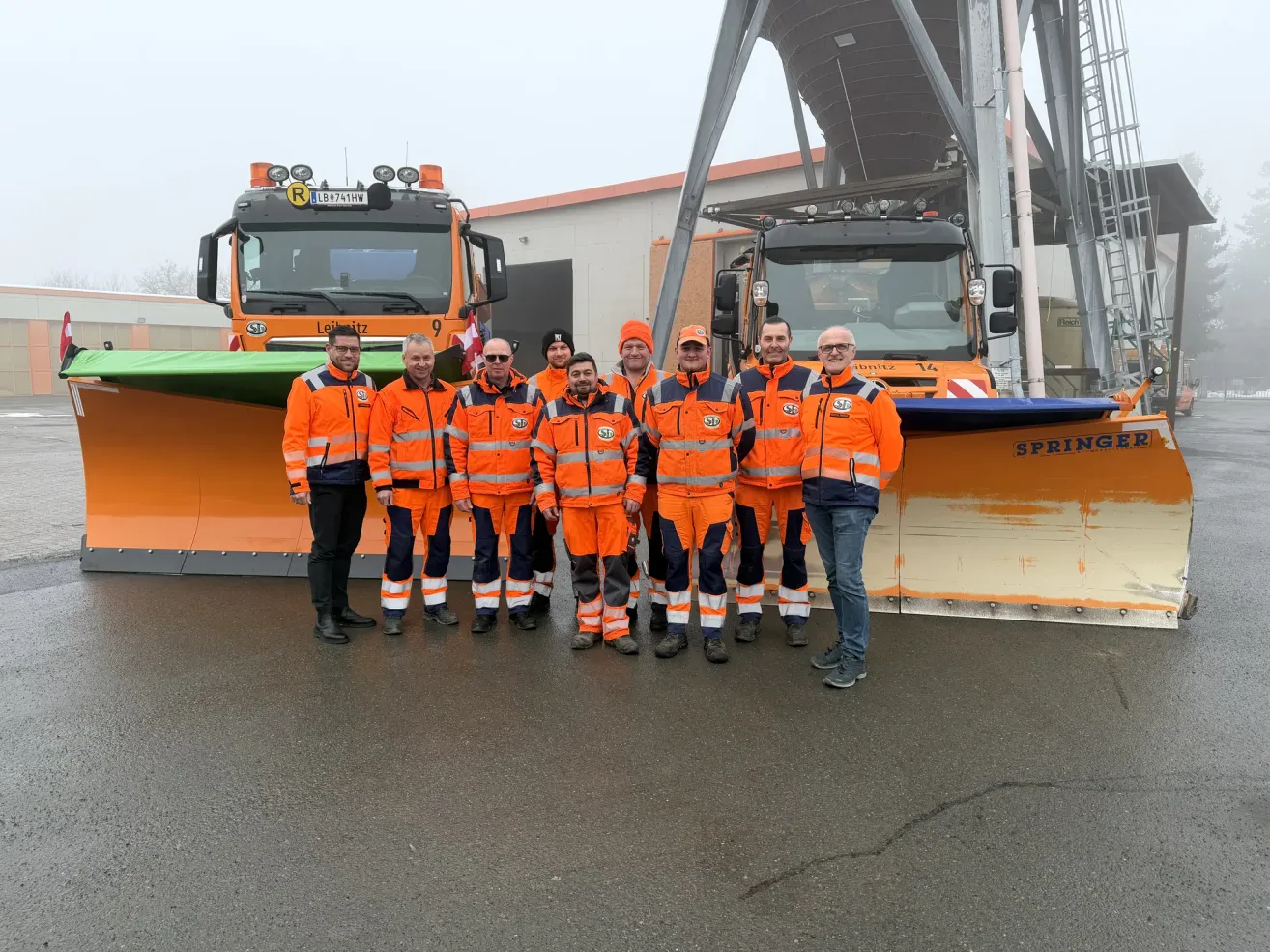 Eine Gruppe von Männern in orangefarbenen Sicherheitsanzügen steht vor zwei Schneepflügen. Einer der Pflüge hat das Wort 'Spring' darauf. Sie lächeln und scheinen für ein Foto zu posieren. Dahinter befindet sich ein Gebäude mit einer grauen Struktur.
