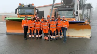 Eine Gruppe von Männern in orangefarbenen Sicherheitsanzügen steht vor zwei Schneepflügen. Einer der Pflüge hat das Wort 'Spring' darauf. Sie lächeln und scheinen für ein Foto zu posieren. Dahinter befindet sich ein Gebäude mit einer grauen Struktur.