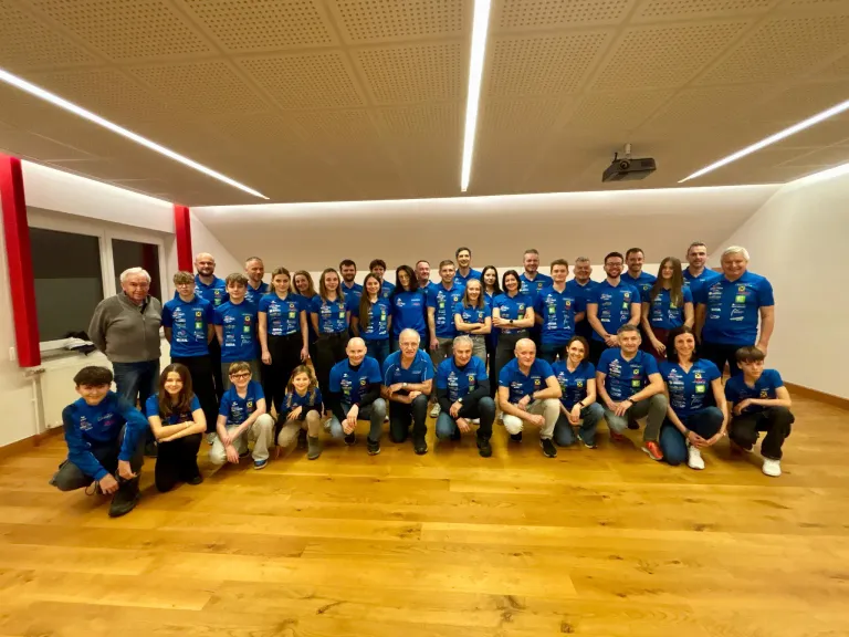 Eine Gruppe von Menschen in blauen T-Shirts posiert für ein Foto in einem Raum mit Holzboden und einer an der Decke hängenden Projektor.