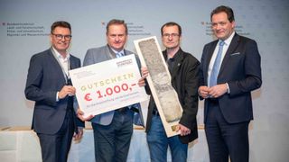 Vier Männer in Anzügen stehen nebeneinander und halten ein Zertifikat und ein Fossil. Das Zertifikat enthält die Wörter 'Erdreich' und 'Gutschein'. Das Fossil ist in einem Holzrahmen ausgestellt. Der Mann rechts hält das Fossil, während der Mann links das Zertifikat hält. Der Mann in der Mitte lächelt. Hinter ihnen ist eine graue Wand mit etwas Text.