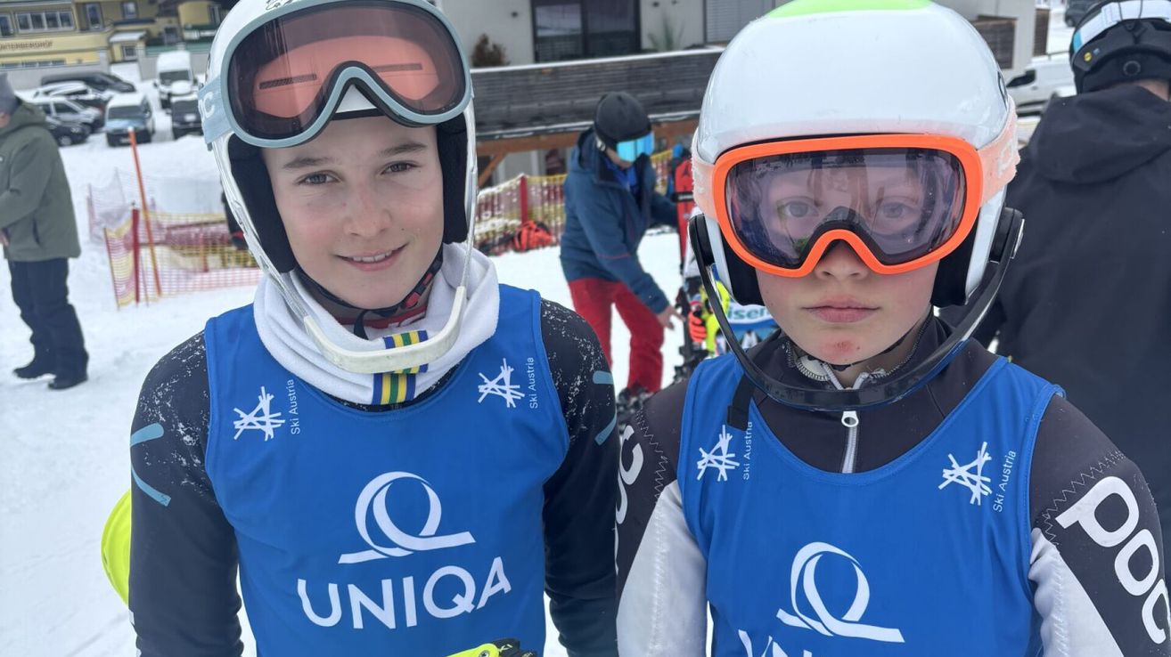 Zwei junge Skifahrer stehen im Schnee und tragen blaue UNIQA-Trikots und Helme. Ein Mann in einer Maske steht hinter ihnen.
