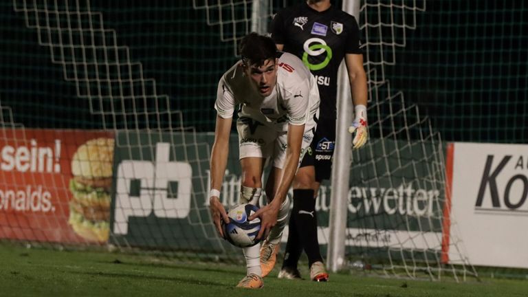 Ein Fußballspieler in weißer Uniform holt einen Ball aus dem Tornetz. Dahinter steht ein weiterer Spieler in schwarzer Uniform nahe des Netzes.