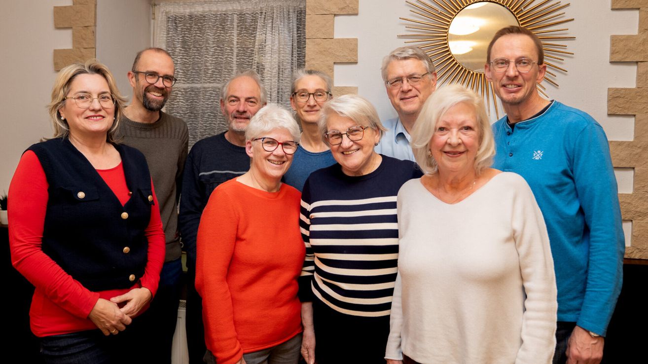 Eine Gruppe von Menschen steht lächelnd zusammen für ein Foto. Hinter ihnen ist eine Wand mit einem Spiegel und einem Fenster mit Vorhängen.