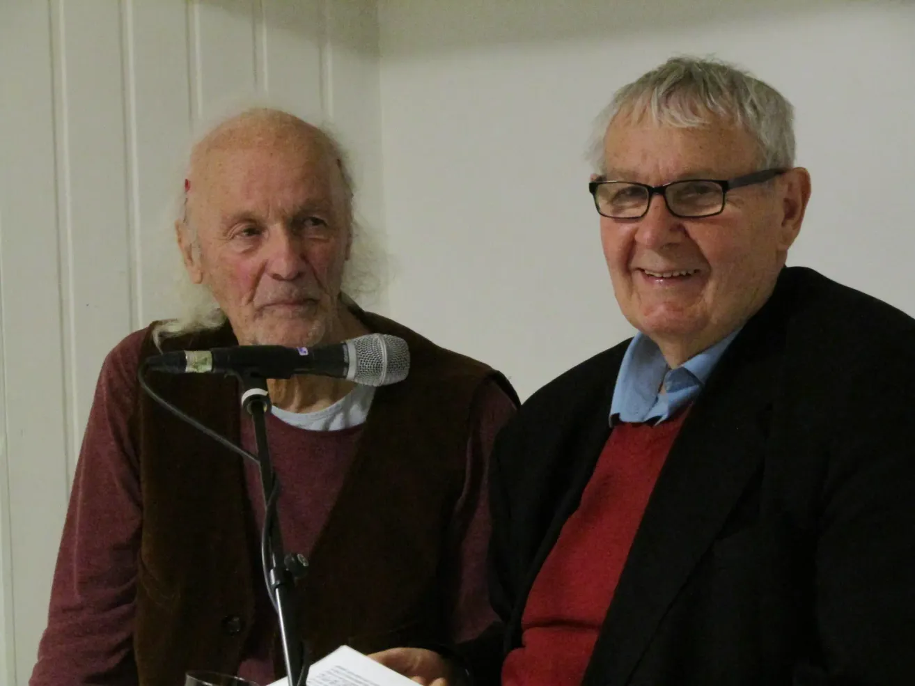 Zwei ältere Männer sitzen nebeneinander, einer mit Brille und der andere mit einem Mikrofon. Sie lächeln beide und scheinen sich bei einer öffentlichen Veranstaltung zu befinden.