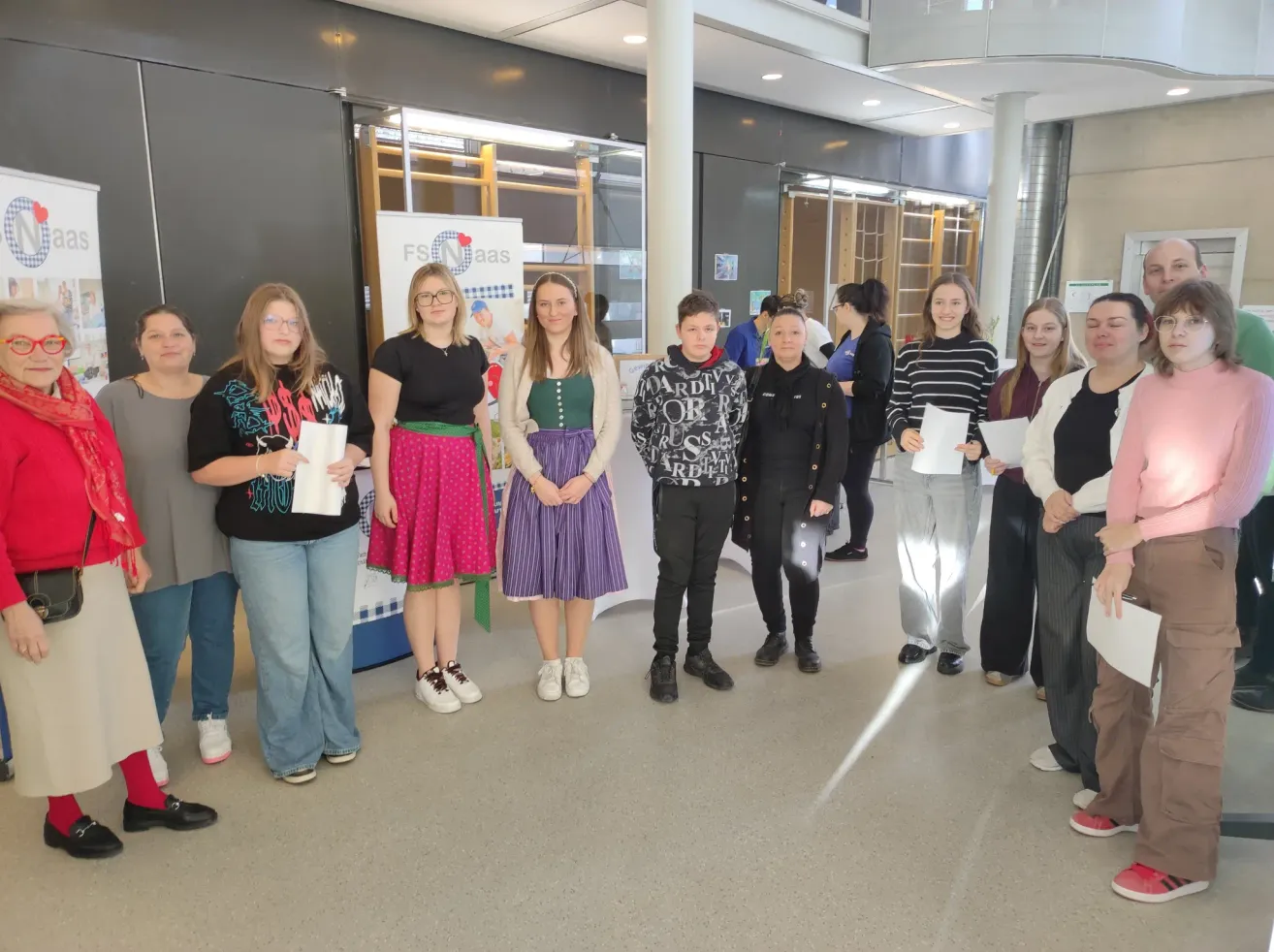 Eine Gruppe von Frauen steht in einer Reihe, hält Papiere in der Hand, trägt verschiedene Kleidungsstücke und Schuhe. Hinter ihnen steht ein Banner mit der Aufschrift 'FSE Maas'. Der Raum hat Glastüren, eine Wand mit Postern und eine gut beleuchtete Decke.