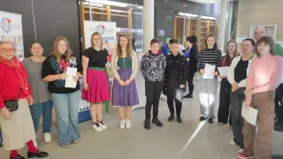 Eine Gruppe von Frauen steht in einer Reihe, hält Papiere in der Hand, trägt verschiedene Kleidungsstücke und Schuhe. Hinter ihnen steht ein Banner mit der Aufschrift 'FSE Maas'. Der Raum hat Glastüren, eine Wand mit Postern und eine gut beleuchtete Decke.