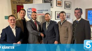 Fünf Männer stehen zusammen, zwei von ihnen schütteln Hände. Sie sind formell gekleidet, wahrscheinlich für ein wichtiges Ereignis. Hinter ihnen ist ein Banner mit dem Text 'Freiheitliche Jugend Kärnten'.