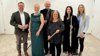 Eine Gruppe von Musikern steht in formeller Kleidung zusammen und hält jeweils ein Musikinstrument. Eine Frau in einem grünen Kleid und ein Mann mit Bart stehen in der Mitte, mit zwei Frauen und einem Mann, die Flöten und eine Klarinette auf beiden Seiten halten.