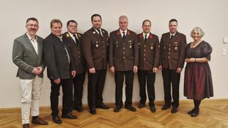 Sechs Männer in Uniformen stehen in einer Reihe, wahrscheinlich in einer formellen Umgebung. Sie tragen Medaillen und Jacken im Militärstil. Hinter ihnen befindet sich eine schlichte weiße Wand.