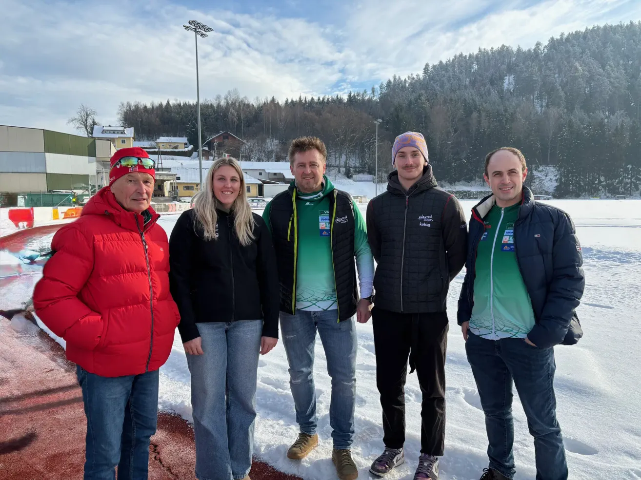 Fünf Personen stehen im Schnee und lächeln für ein Foto. Hinter ihnen sind Berge und ein bewölkter Himmel zu sehen. Zwei von ihnen tragen Mützen. Sie sind für kaltes Wetter gekleidet.